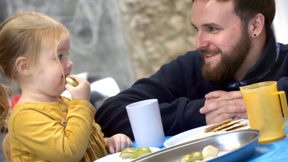 Little girl in a yellow dress enjoying a snack, engaging with a smiling bearded man during mealtime.