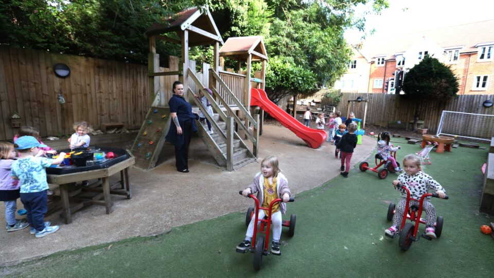 Children playing outside at a pre-school, riding tricycles and using a climbing frame with a red slide.