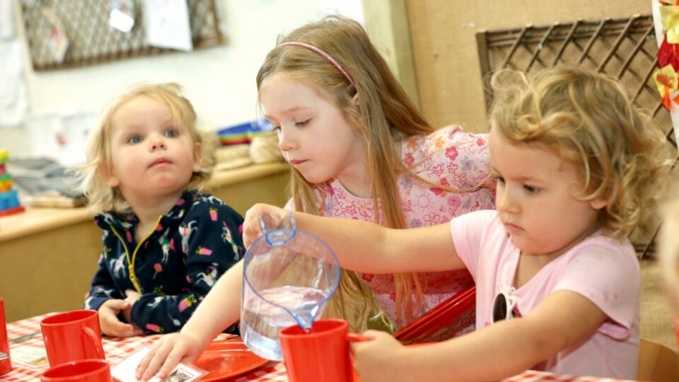 Nursery children sit around a table, as one girl carefully pours water into a cup during snack time.