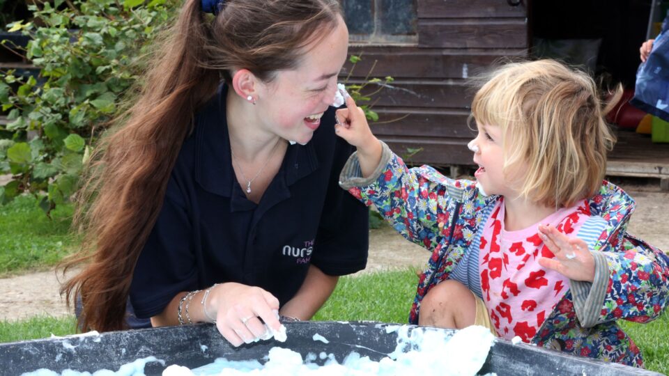 A smiling nursery worker and a young child playing with foam, as the child playfully touches her face.