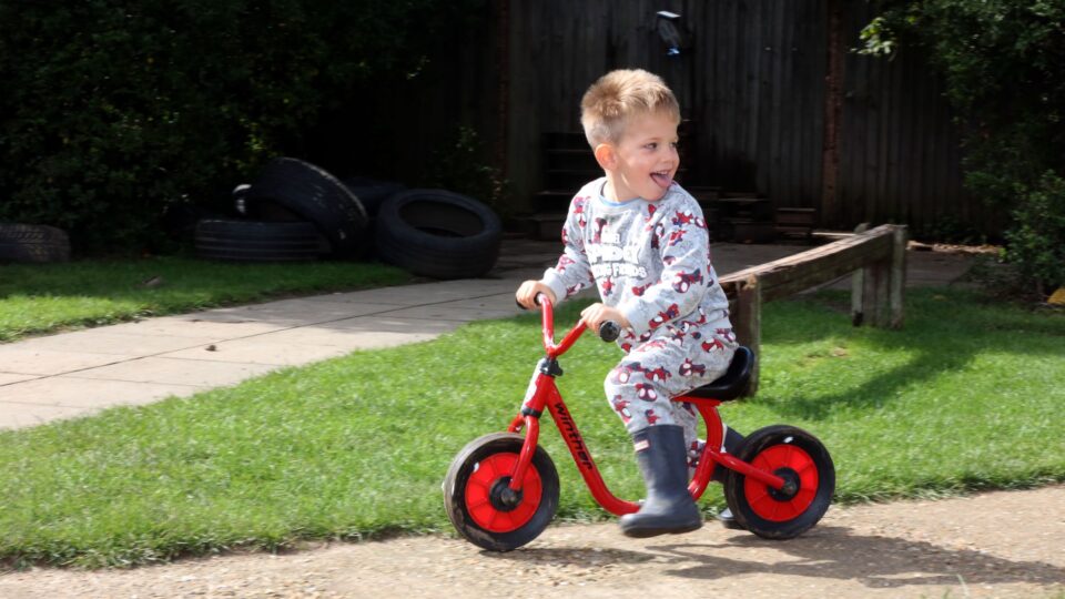 A young boy rides a red balance bike outdoors, smiling as he enjoys playtime at the nursery.