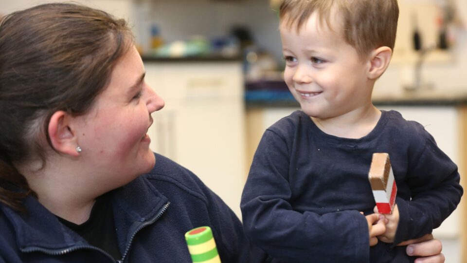 A caregiver at a day nursery in West Sussex smiles warmly at a young boy holding a wooden toy.