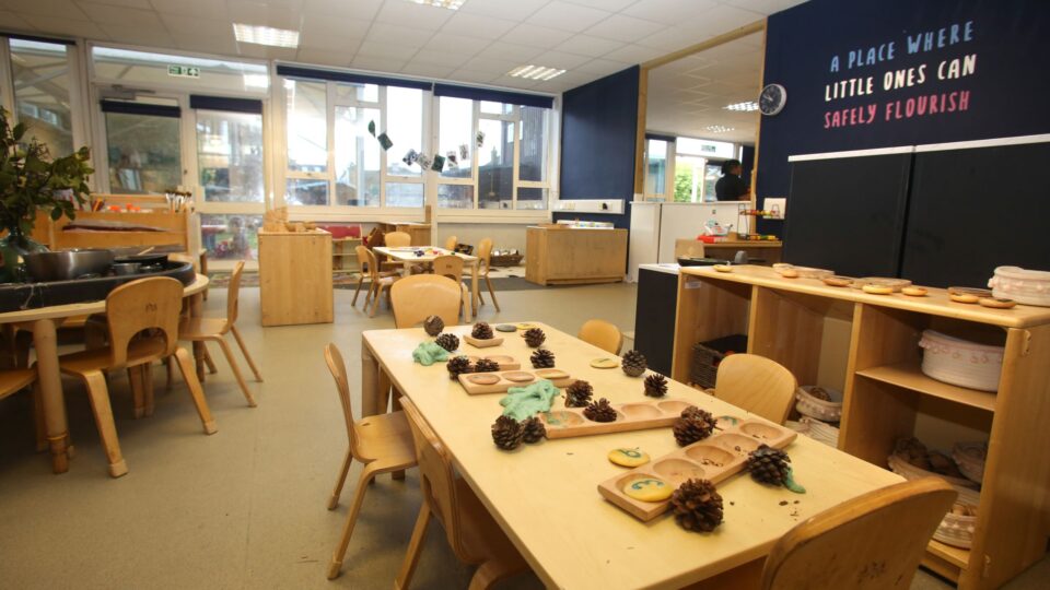 Shoreham day nursery classroom with wooden tables, pinecones, and educational materials, under a sign that reads "A place where little ones can safely flourish."