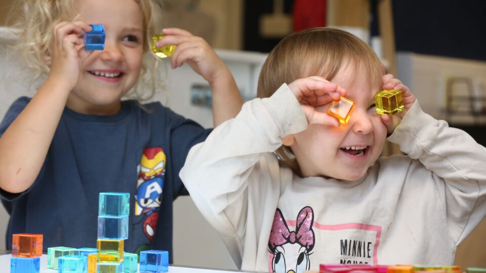 Smiling children enjoying playful learning with colourful translucent blocks, stacking and holding them to their faces.