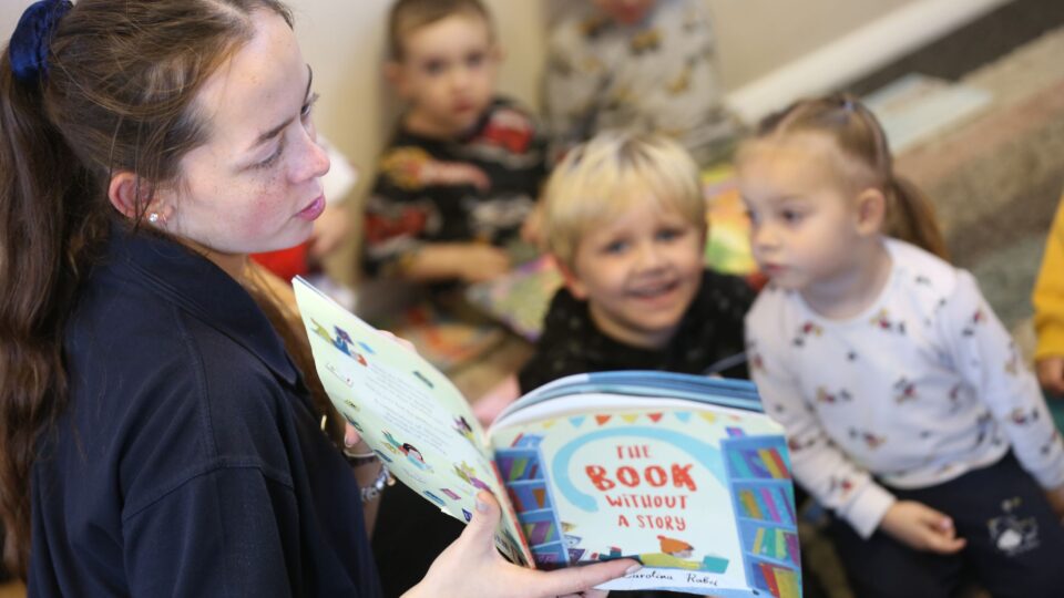 A nursery worker reads "The Book Without a Story" to a group of attentive children during storytime.