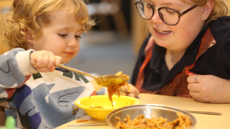 A toddler uses a spoon to serve food into a bowl while a nursery worker watches with a smile.