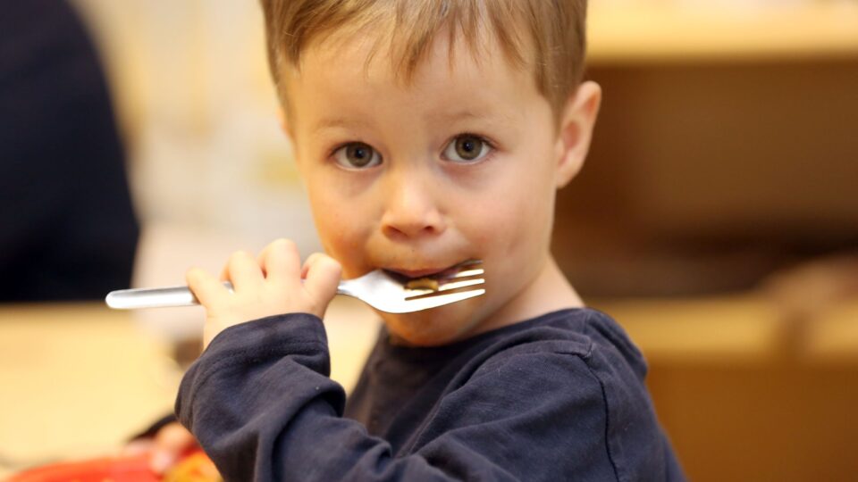 A young boy eating with a fork looks directly at the camera while enjoying his meal.