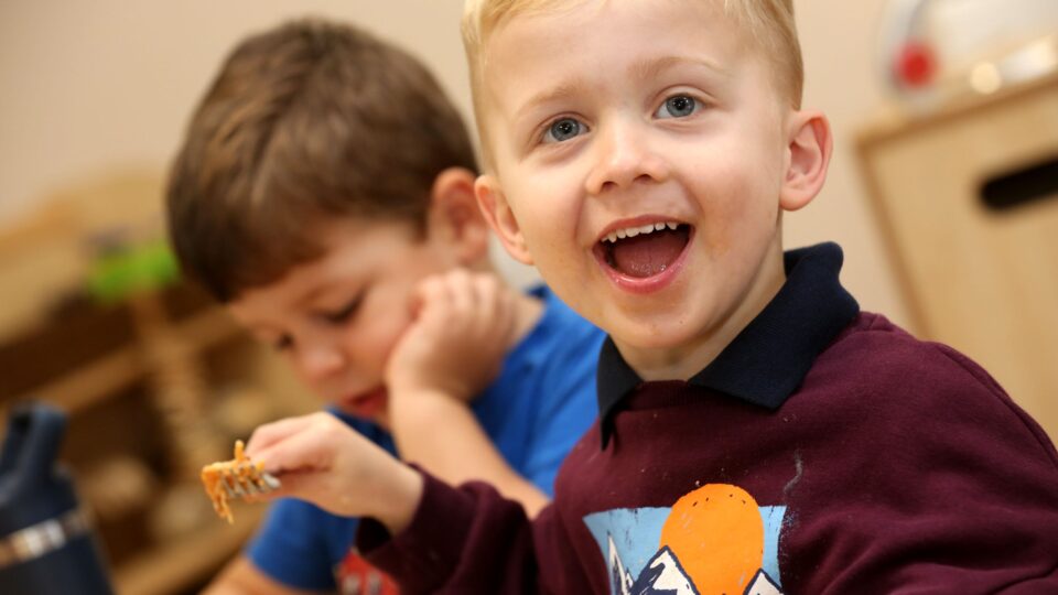 A smiling boy enjoys his meal at nursery while another child focuses on eating in the background.