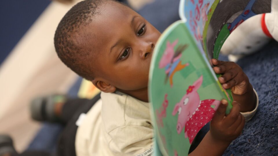 A young boy deeply focused on reading a colourful book as part of his nursery education.