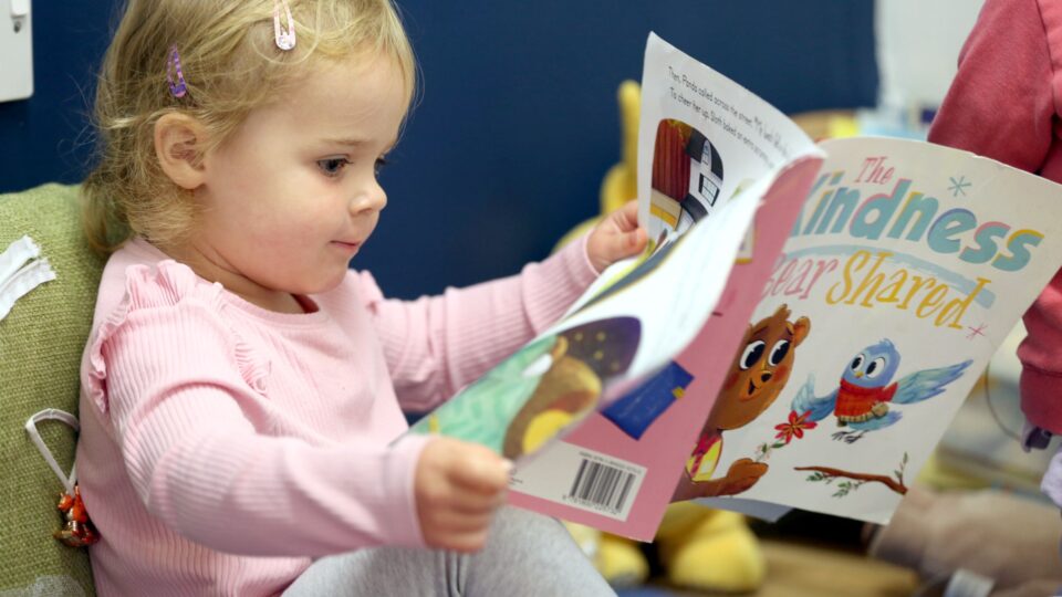 A toddler in a day nursery intently reads a colourful picture book, focused on the story, illustrating early literacy development.